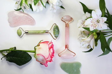 Jade face roller for beauty facial massage therapy. Beauty tools pink and green face roller  on flowers background. Face treatment. Home spa.  