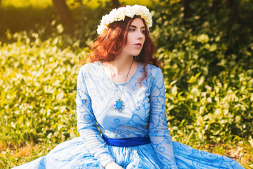 Portrait of an attractive woman with a rim of white flowers and in a blue magnificent dress sitting on the grass in the park against the background of lilac bushes. 