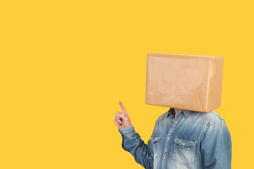 Hombre con caja de cartón en la cabeza y con un gesto con el dedo señalando hacia arriba sobre fondo amarillo liso brillante aislado. Vista de frente. Copy space
