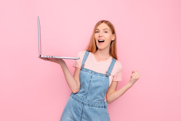 Portrait of a shocked girl holding a laptop and showing a winning gesture isolated on a pink background