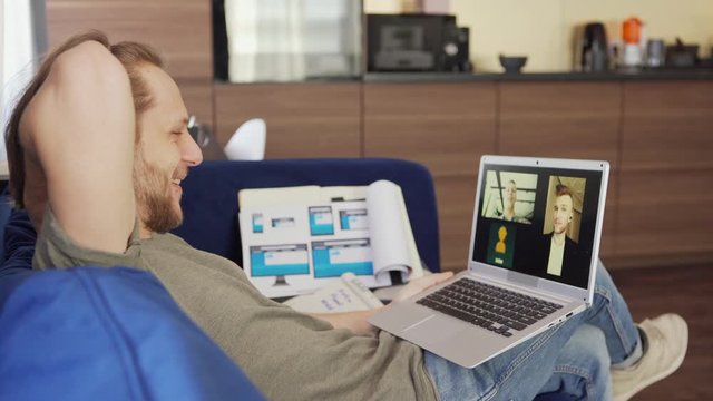 Side view of young businessman having video conference with two colleagues on laptop computer sitting on sofa at home