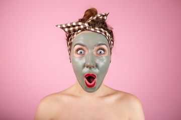 Portrait of a girl with red in a clay mask on her face standing on a pink background, shaken, degenerated, surprised emotions on her face