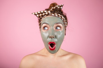 Portrait of a girl in a clay mask on her face standing on a pink background expressing surprised emotions on her face