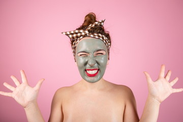 Girl in a clay mask on her face standing on a pink background, shaken by a burning skin from the mask. Satisfied girl on a skin care procedure.