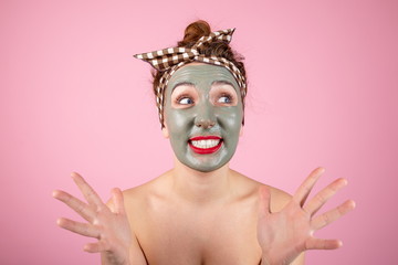 Portrait of a girl in a clay mask on her face standing on a pink background, scared by the burning skin of the mask. Satisfied girl on a skin care procedure