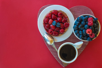 Bowl with blackberries, cake with red raspberries, cup of coffee on board shape heart on red background. Space for text, top view