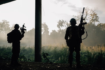Two military armed soldiers standing in a building with rifles look towards a dense forest. War...