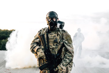 Armed soldier in a gas mask with a rifle standing on the roof of a building in smoke. Strona...