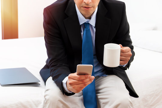 Shot of a handsome businessman sitting on the bed with smart phone and coffee