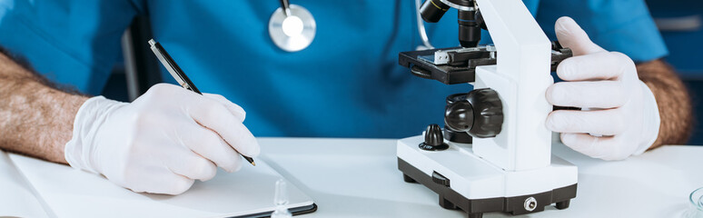 Fototapeta na wymiar partial view of biologist in latex gloves writing in notebook near microscope, panoramic shot