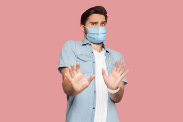 Fototapeta na wymiar Stop. Portrait of scared man with surgical medical mask in blue shirt standing with afraid or worry face, looking at camera and blocking with his hands. indoor studio shot, isolated on pink background