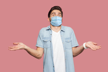 I don't know. Portrait of confused man with surgical medical mask in blue casual style shirt standing with raised arms, looking at camera with answer. indoor studio shot, isolated on pink background.