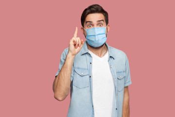 I have an idea. Portrait of surprised young man with surgical medical mask in blue shirt standing with amazed face, looking at camera with idea sign. indoor studio shot, isolated on pink background