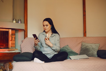 Obraz premium A girl sits on a sofa at home and drinks rose wine against the background of the fireplace. Stay at home, the girl is resting in the evening, relaxation. The girl is clueless writes SMS. Girl drinks 