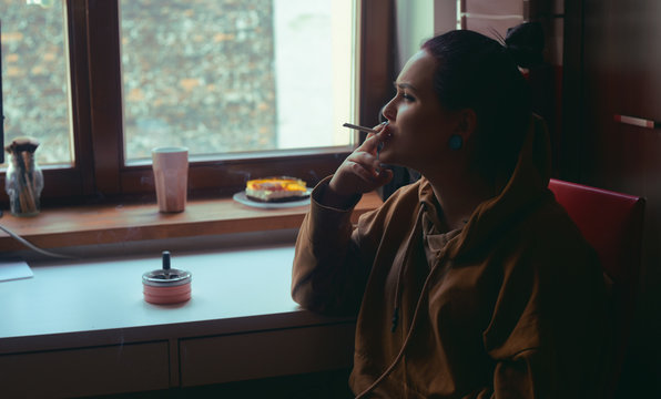 The girl at home sits and smokes, looks out the window. Lonely girl smokes by the window.