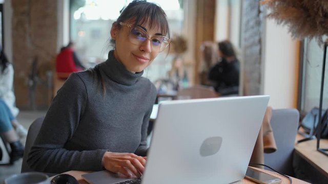 Portrait young woman in eyeglasses using laptop in coworking space or cafe and smiling to camera. female sitting in front open laptop computer. Study, learning, remote work, freelance.
