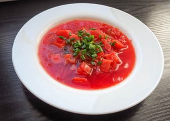White round plate with soup of national cuisine borscht with sliced green onion rings on the background of a dark wooden table