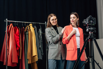 selective focus of stylist in glasses touching trendy jacket on attractive model near digital camera