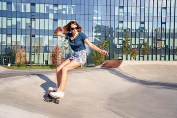 Young woman in short skirt skating on longboard outdoor in a skate park, doing the trick. Focus on background.