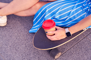 Skater woman in dress is sitting on longboard with paper cup of coffee. Close up view on hand and board.