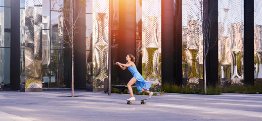 Skater woman in dress is training on longboard, doing a trick outdoor in a public park.