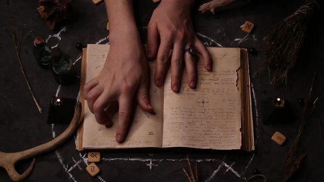 Top view of a female witch fortune teller reading, leading her hands through the pages of an ancient book in an unknown language. Magic black candles on the background of the pentagram.