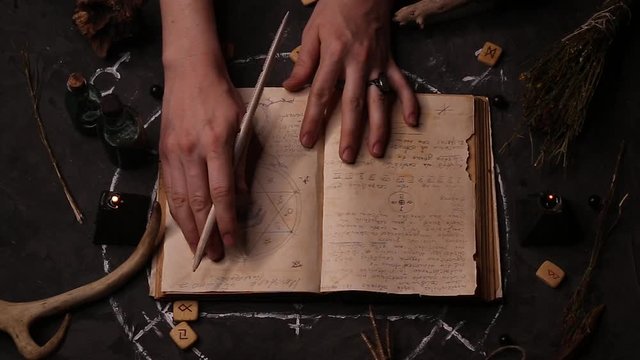 Top view of a female witch fortune teller reading, leading her hands through the pages of an ancient book in an unknown language. Magic black candles on the background of the pentagram.