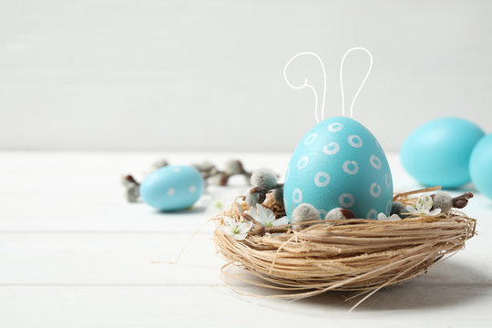 Colorful Easter egg with cute bunny ears in decorative nest on white wooden table. Space for text