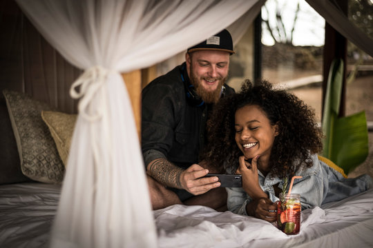 Happy couple on bed in a lodge with smartphone and fresh ice tea