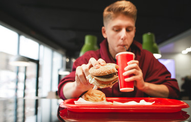 Tray with fast food, french fries, burger in the hands of a young man drinking a drink from a red...