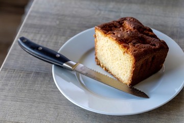A portrait of a apple pound cake on a white plate on a wooden table with a bread knife on the plate. It is made from apples, sugar, flour, eggs and butter and is baked in an oven.