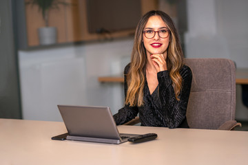 A bespectacled businesswoman on a computer looks at the camera with a smile.