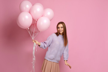 Smiling carefree girl in knitted sweater dancing with pastel pink air balloons isolated on pink background. Beautiful happy young woman on a birthday holiday. space for text