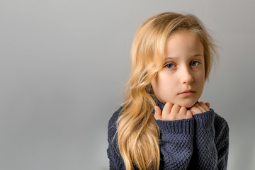 Color portrait of serios blond child girl, headshot portrait of sad little girl in grey pullover posing in studio on the grey background