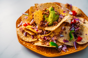 Close up of a plate of delicious Tortilla nachos corn chips with melted cheese sauce, ground beef, jalapeno peppers, red onion, salsa, and sour cream with guacamole dip. Top view