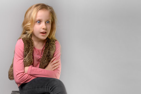 portrait of surprised blond child girl of 8 years old, portrait of emotional little kid in pink T-shirt and fur vest in studio on the grey background, copy space