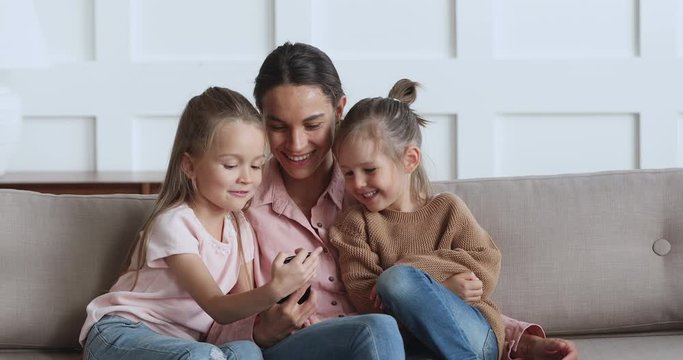 Young pretty woman sitting on sofa with cute smaller sisters, showing funny mobile application. Happy little adorable children girls siblings laughing at cartoon photos video on smartphone with mum.