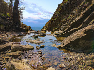 A stream flowing past the rocky mountains and flowing into the sea on a sunny day with a blue cloudy sky.