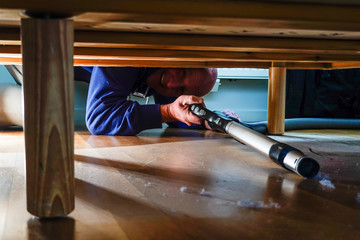 A man vacuums dust under a bed in an apartment