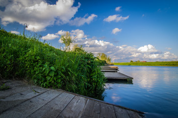 Wide angle view of a waterfront wooden pier surrounded by green vegetation, under a blue sky with...