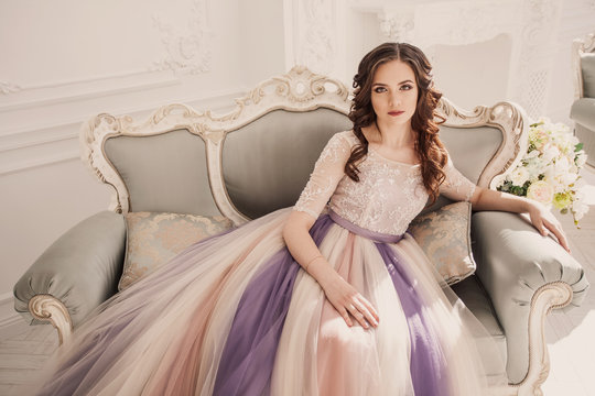 Curly girl in an original dress sits on a sofa. Beautiful model in a beautiful wedding dress in a white photo studio. Bride in color wedding dress.