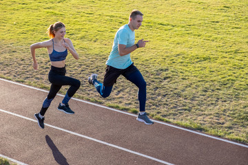 Young couple of fit sportsmen boy and girl running while doing exercise on red tracks of public stadium outdoors.