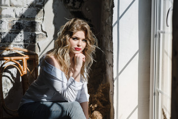 Young woman sitting in front of window. Girl sitting on a windowsill. The young blonde girl is sitting at the window in jeans and a white shirt. Photography with beautiful girl