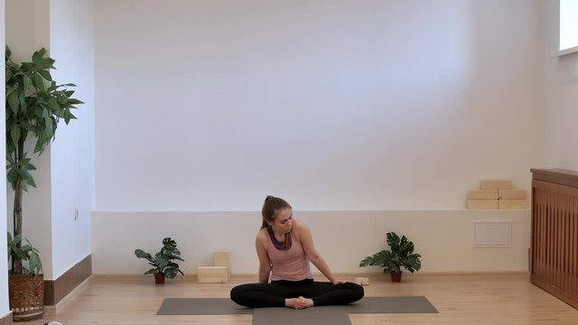 A yoga teacher in a Pilates studio on a yoga mat performs a deep opening of the hip joints in a butterfly pose on her left foot. Baddha Konasana.