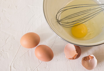 one broken egg in a Cup with a whisk and two whole eggs with a shell on a white surface
