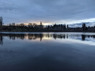 Fototapeta premium Deep blue dusky sky over a lake, with lightly wooded area across the lake in the background. Cool colors visible in both the water and the sky except for a warm area near the horizon.