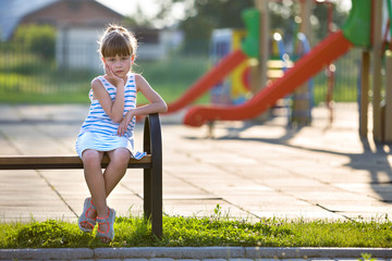 Fototapeta na wymiar Cute young girl in short dress sitting alone outdoors on playground bench on sunny summer day.