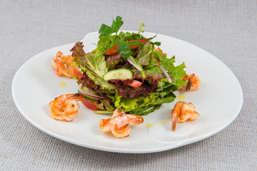 Salad of lettuce, tomato, cucumber, purple onion with lemon oil decorated with king prawns on a round original plate on a gray background