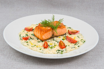 A large piece of fried salmon with rice porridge and butter, decorated with greens and tomatoes, as well as dill on a large round plate on a gray background