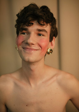 Portrait of a queer guy with curly hair and golden earrings who is smiling topless